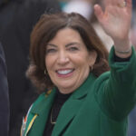 A smiling woman in a green coat waving at an outdoor event