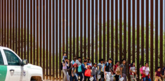 Group of families and children walking near a border wall with a customs vehicle nearby