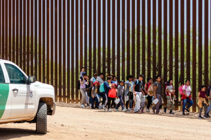 Group of families and children walking near a border wall with a customs vehicle nearby
