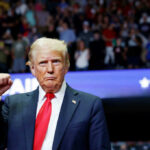 A man in a suit with a red tie raises his fist at a rally, with a cheering crowd in the background
