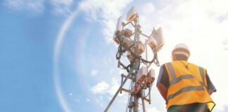 Telecommunication worker in safety gear standing near a cell tower under a blue sky