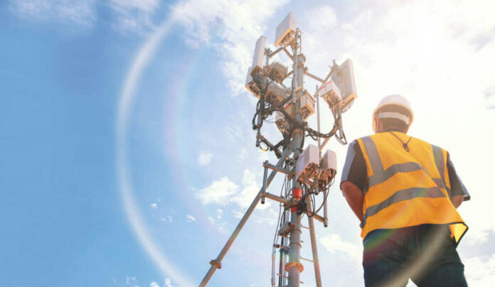 Telecommunication worker in safety gear standing near a cell tower under a blue sky