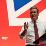 A man in a white shirt and tie gestures while speaking at a podium with a British flag backdrop