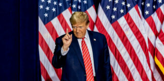 A man in a suit gesturing with one finger in front of multiple American flags