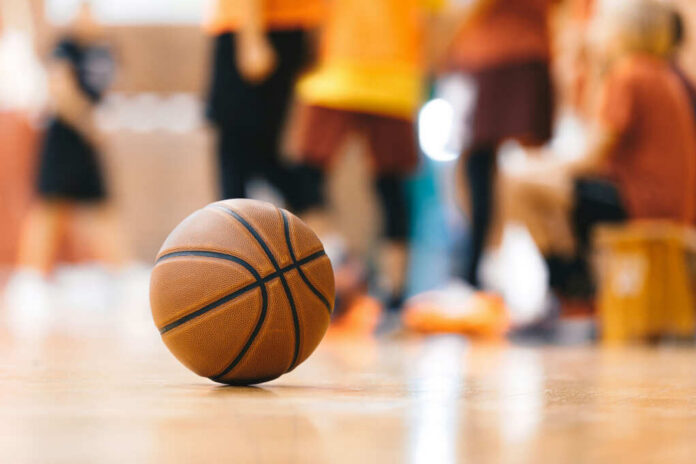 A basketball resting on the gym floor with players blurred in the background