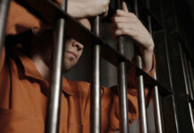 A male prisoner in an orange jumpsuit holding onto the bars of a jail cell