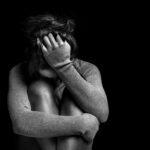A person sitting in a curled position, covering their face with one hand in a dark setting