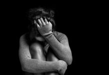 A person sitting in a curled position, covering their face with one hand in a dark setting