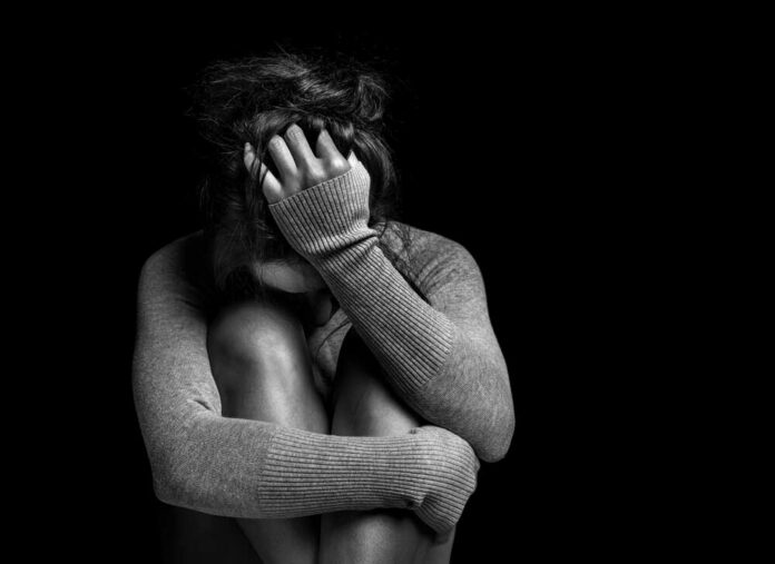 A person sitting in a curled position, covering their face with one hand in a dark setting