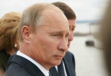 Close-up of a serious-looking man in a suit at an outdoor event near water