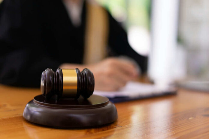 shutterstock_2224687169.jpg A gavel on a wooden table with a blurred figure of a person in professional attire in the background