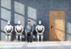 A man sitting between three humanoid robots in a waiting room for a job interview