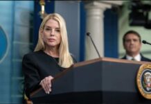 A woman at a podium during a press conference with a man in the background