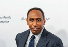 A man in formal attire smiling at a media event with a branded background