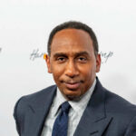 Stephen A. Smith BLASTS Democrats – Unreal! A man in formal attire smiling at a media event with a branded background