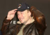 Man in a leather jacket and baseball cap smiling and adjusting his hat