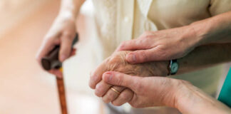 Two hands clasped together, one elderly and one younger, with a cane in the background