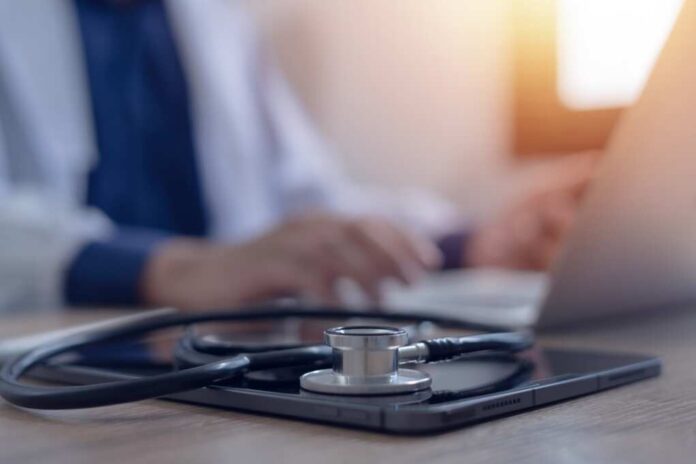A stethoscope resting on a tablet with a blurred medical professional in the background typing on a laptop