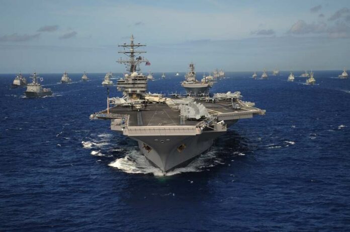 shutterstock_2599523537.jpg An aircraft carrier surrounded by various naval ships in the ocean