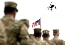 Military personnel standing in formation with a drone flying overhead and an American flag in the background