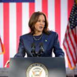 A female political leader speaking at a podium with American flags in the background