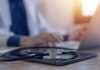 A stethoscope resting on a tablet with a blurred medical professional in the background typing on a laptop