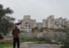 Hebron Horror: Infant Dead, Answers Missing Person holding a Palestinian flag in front of buildings and a barrier