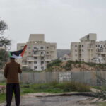 Person holding a Palestinian flag in front of buildings and a barrier