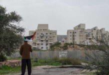 Hebron Horror: Infant Dead, Answers Missing Person holding a Palestinian flag in front of buildings and a barrier