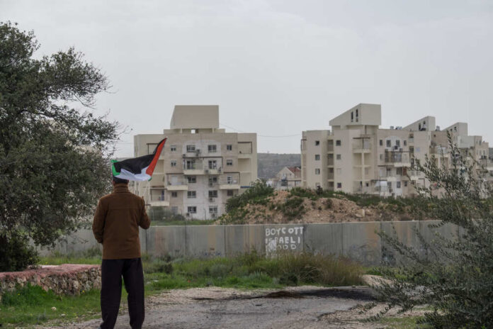 A,Palestinian,Protester,Stands,Near,The,West,Bank,Barrier,Holding Person holding a Palestinian flag in front of buildings and a barrier