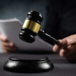 A gavel held above a sounding block with a person reading documents in the background