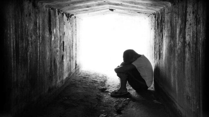 A person sitting in a dark tunnel, hugging their knees with light visible at the end