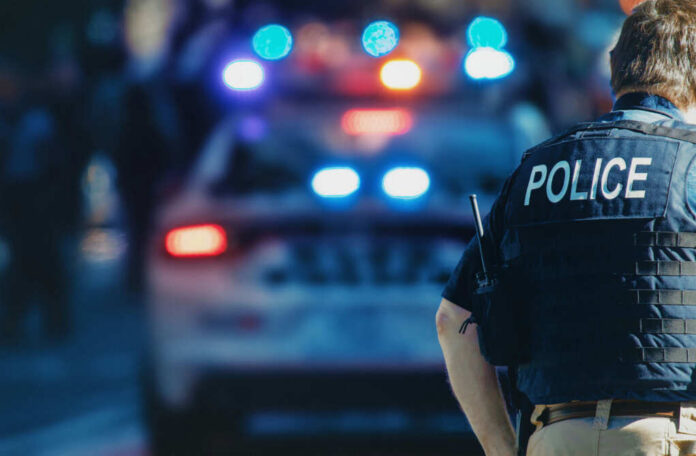 shutterstock_1543591088-1 Police officer in a tactical vest standing in front of a police car with flashing lights