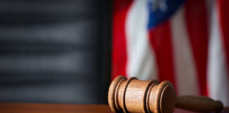 A wooden gavel resting on a round base with a blurred American flag in the background