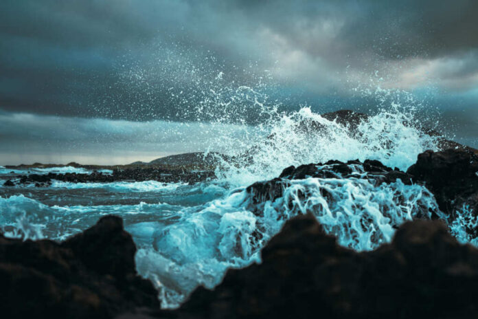 shutterstock_1692249964 Waves crashing against rocks under a cloudy sky