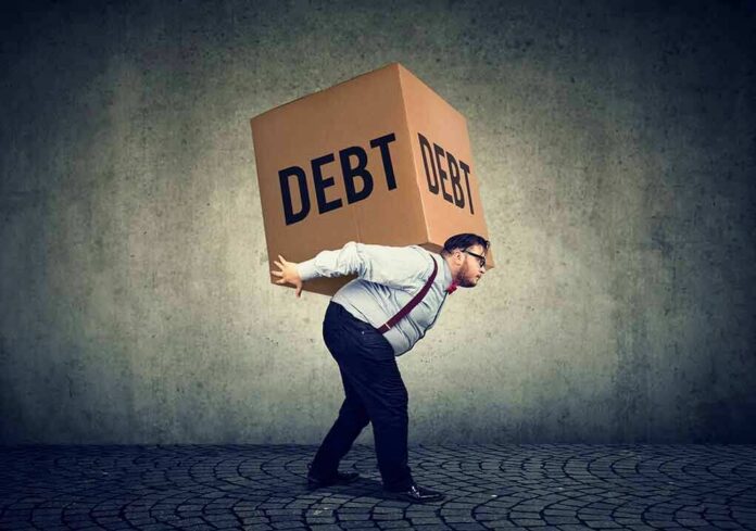 shutterstock_1044442636 Man carrying a large box labeled debt on his back