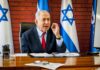 Phased Disarmament Sparks Explosive Standoff man in suit speaking at a desk with Israeli flags behind him