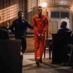 New Mexico Justice Runs On Deals Inmate in orange jumpsuit escorted by officer in courtroom