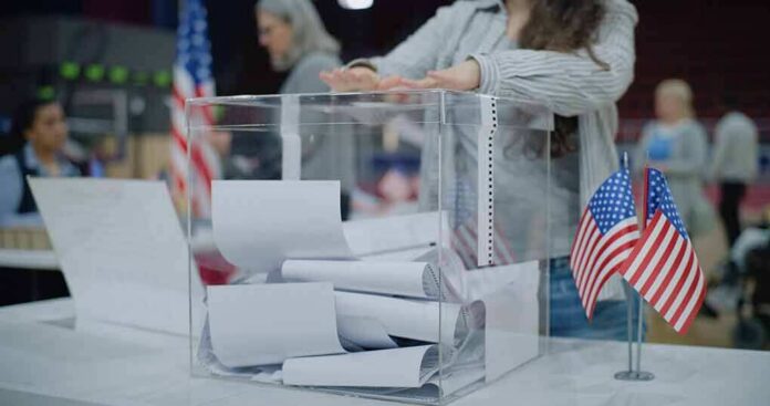 shutterstock_2454758129-1 Person casting a ballot into a clear box at a polling station