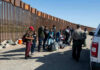 DHS Boast Sparks Data Fight Group of people standing by a border fence with officers and vehicles