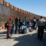 DHS Boast Sparks Data Fight Group of people standing by a border fence with officers and vehicles