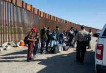 DHS Boast Sparks Data Fight Group of people standing by a border fence with officers and vehicles