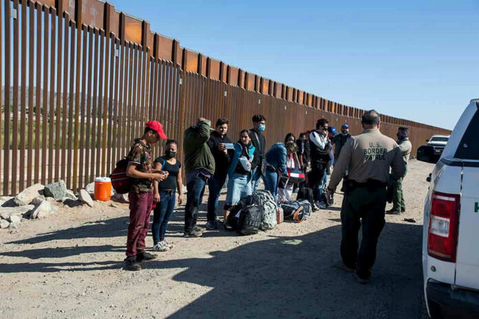 Group of people standing by a border fence with officers and vehicles