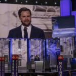 Eligibility Bomb Threatens Maine Frontrunner Man speaking at podium on stage, projection behind him.