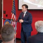 Abuse Firestorm Engulfs Trump-World Ally Speaker in a navy suit addressing an audience at a podium