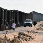 Land Grab Fears Explode in Texas Border fence with patrol vehicles and people walking on a dirt road