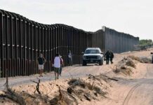 Border fence with patrol vehicles and people walking on a dirt road