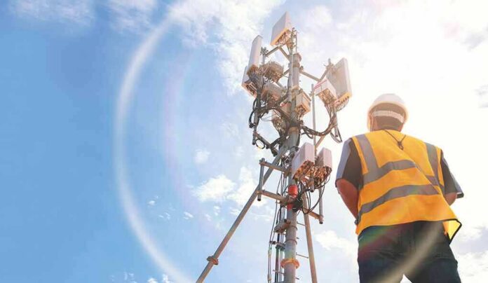 shutterstock_2106490226 Telecom tower with worker in safety vest under bright sky