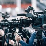 Fetal-Cell Fight Erupts Amid Measles Surge Broadcast cameras on tripods at a press event
