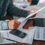 Vance’s $230B Claim — Where’s The Proof? Person reviewing bills with calculator at cluttered desk